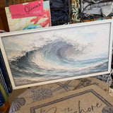 A framed square art print depicting a large ocean wave curling, displayed on a table in a retail setting. In front of the frame is a beige doormat with the script text 'by the Seashore' and nautical motifs.