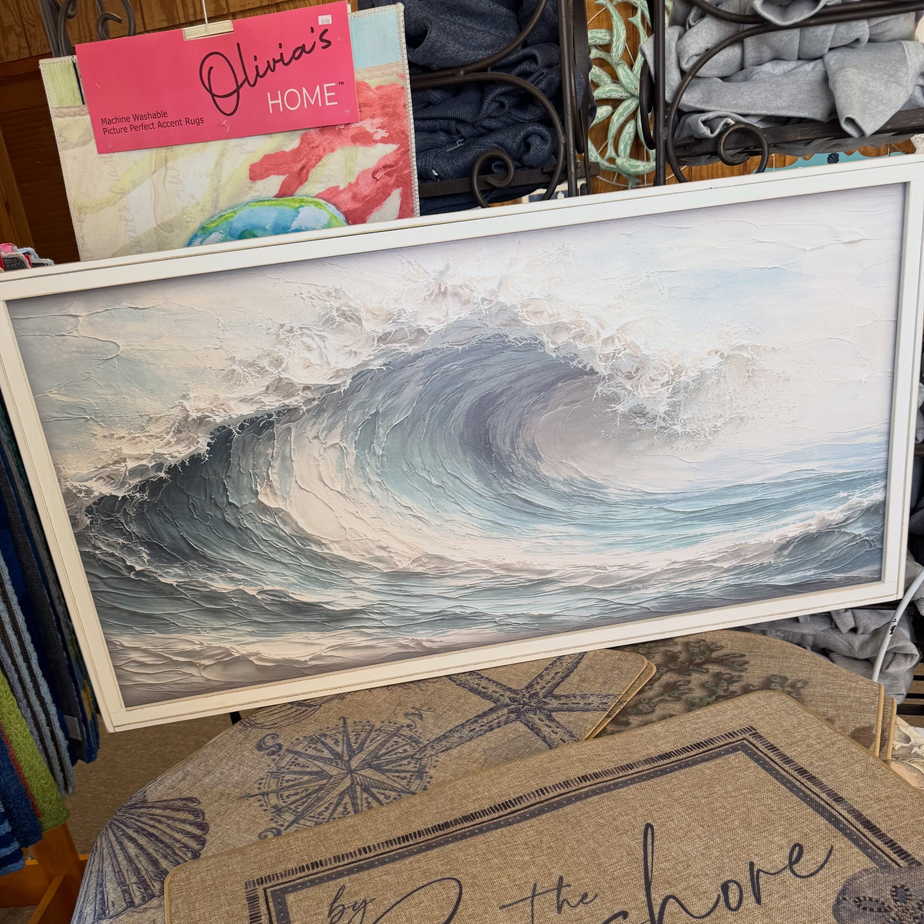 A framed square art print depicting a large ocean wave curling, displayed on a table in a retail setting. In front of the frame is a beige doormat with the script text 'by the Seashore' and nautical motifs.