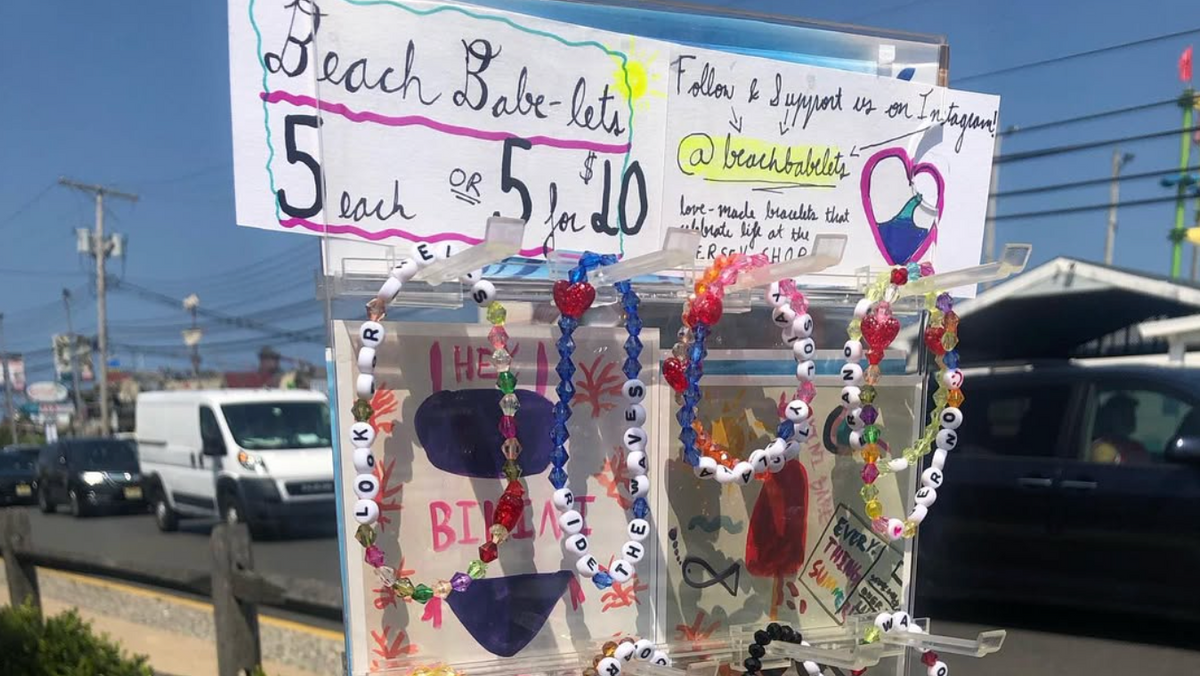 A clear acrylic jewelry display stand filled with colorful beaded bracelets at an outdoor market. Handwritten signs above the stand advertise 'Beach Babe bracelets' with pricing, and a street scene with cars, bushes, and a person's leg is visible in the background.